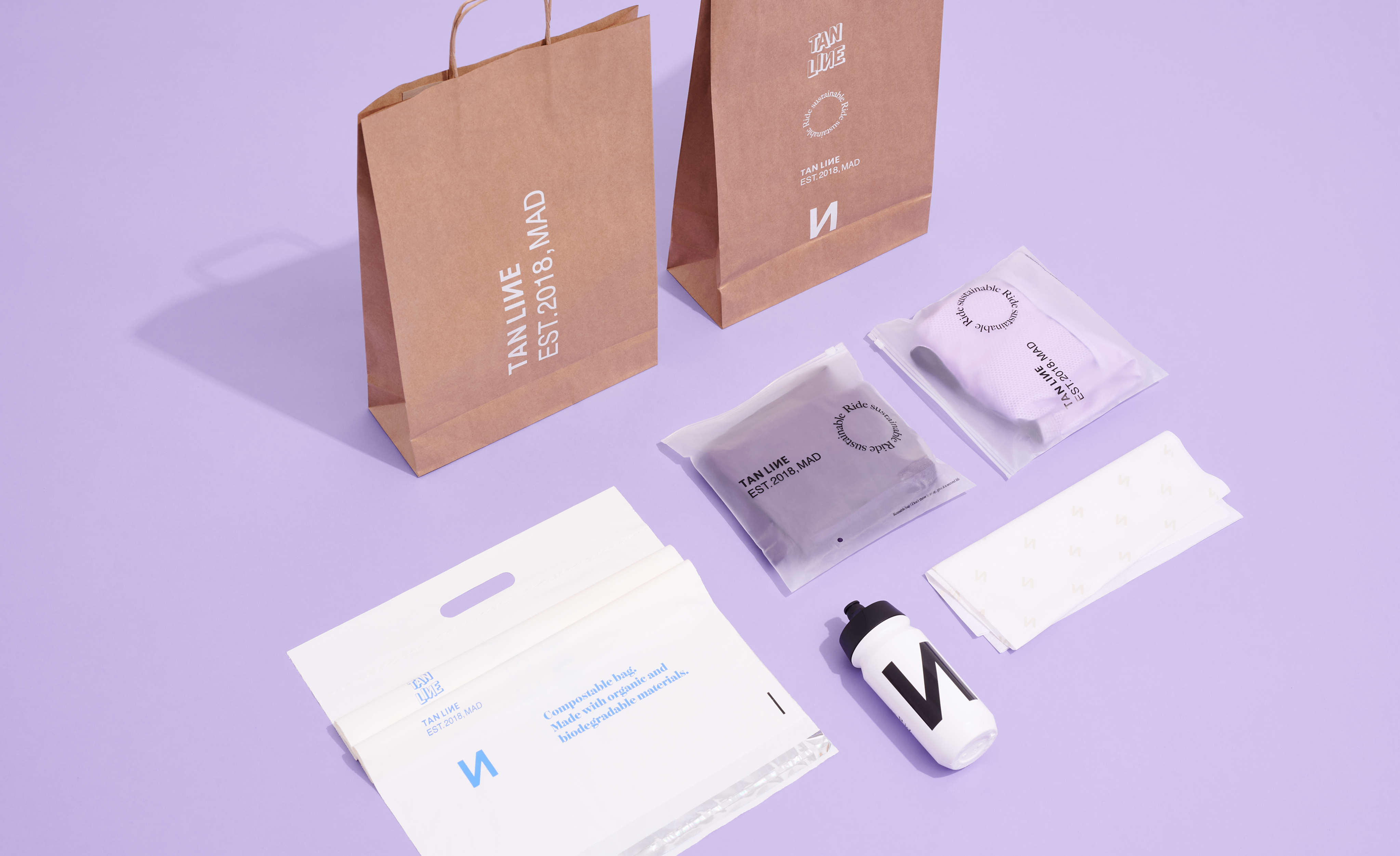 Branded paper shopping bags, clothing pouches, mailing bags, tissue paper, and a reusable bottle arranged on a purple background